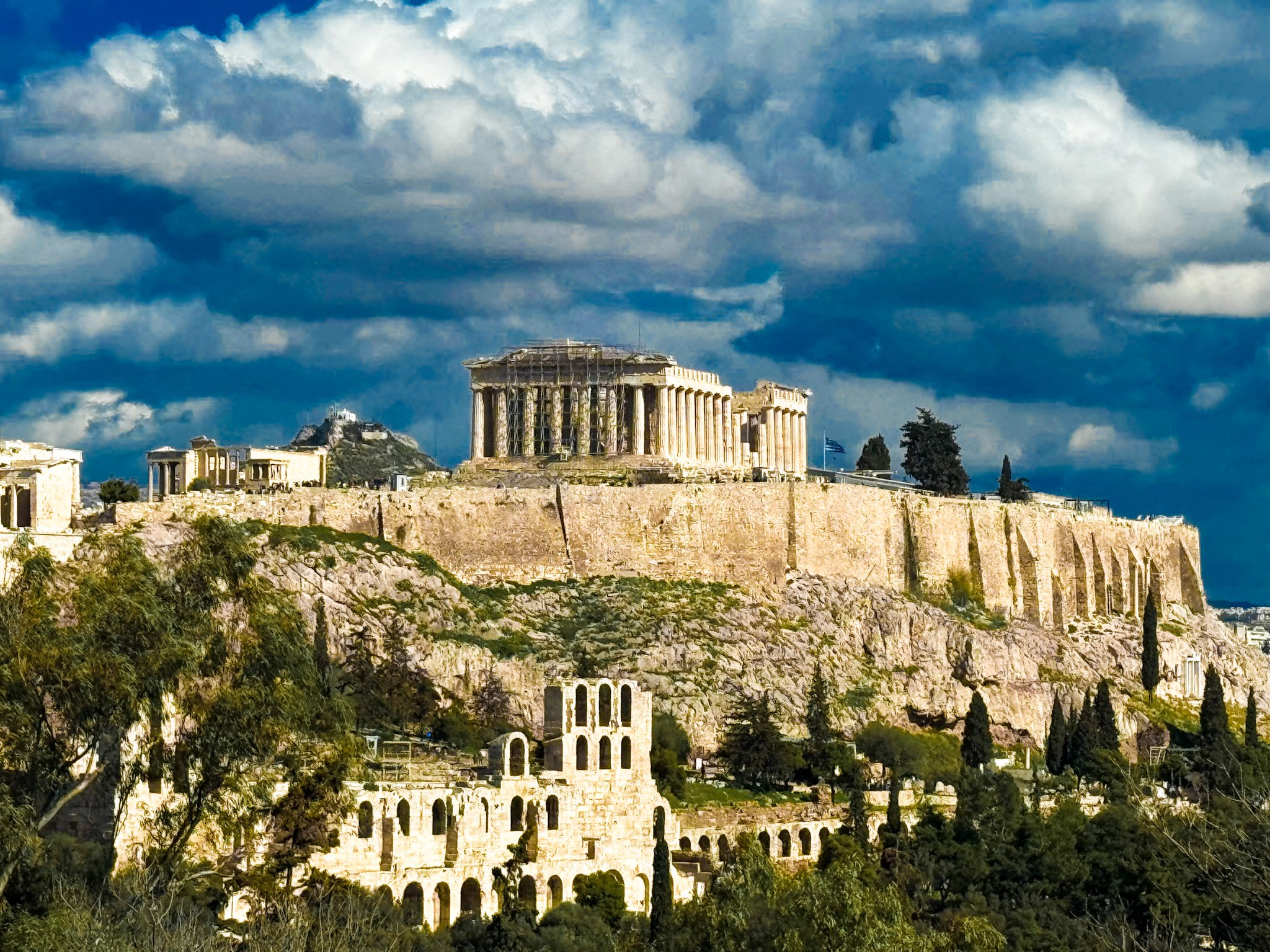 Klasyczny widok na Akropol z Partenonem i Odeonem Heroda Attyka w Atenach. Imponujące mury starożytnej twierdzy na skalistym wzgórzu, w tle widoczna flaga Grecji i Wzgórze Lycabettus pod kłębiastymi, ciemnymi chmurami.