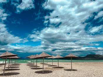 Straw umbrellas casting shadows on a pebble beach in Corinthia, Greece, with clear turquoise waters and distant mountains under a dramatic sky, ideal for a day trip from Athens.