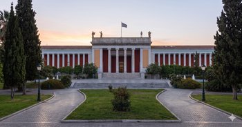Monumentalny budynek Narodowego Muzeum Archeologicznego w Atenach o wschodzie słońca, z kolumnami, posągami i grecką flagą, otoczony zielonym parkiem. Muzeum to jedna z głównych atrakcji Grecji, prezentująca skarby antyku, takie jak Maska Agamemnona i mechanizm z Antykithiry.