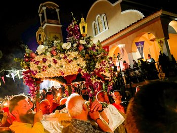 Nighttime Easter procession in Greece in front of a traditional church. A group of men carry the Epitafios on their shoulders - a flowered bier representing the tomb of Christ, decorated with white and purple flowers and topped with golden lanterns. A crowd of faithful holding lit candles is visible in the background.