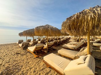 Rows of empty sunbeds with beige cushions and straw umbrellas on a sandy beach, facing the calm blue sea under a clear sky, illustrating a premium beach club experience in Athens, Greece, where advanced reservations are recommended for 2026.