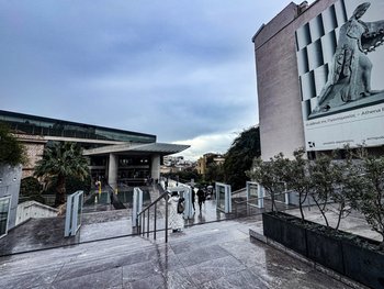View of the modern entrance to the Acropolis Museum in Athens, featuring glass architecture and a large banner of the goddess Athena beside the main stairway.