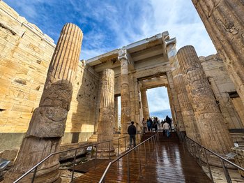 Tourists walk on a wooden ramp through the Propylaea, the monumental gateway to the Acropolis of Athens, featuring ancient fluted columns and stone architecture under a blue sky. This image highlights the entrance experience for visitors exploring Acropolis tickets and the historic archaeological site in Athens.