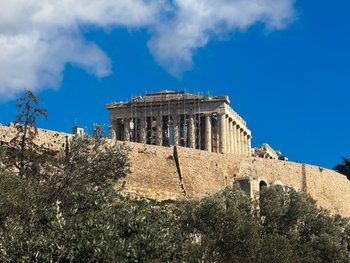 View of the Acropolis from Philopappos Hill in Athens, featuring the Parthenon and Odeon of Herodes Atticus under a dramatic sky with olive trees in the foreground.