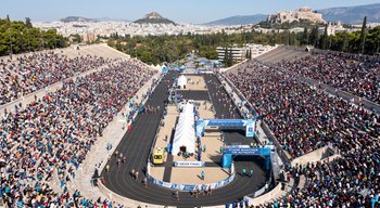 Aerial view of the Panathenaic Stadium in Athens, Greece, filled with spectators cheering runners crossing the finish line of the Athens Classic Marathon, with the Acropolis visible in the background. This iconic finish line is a key part of the Athens Marathon 2026 experience.