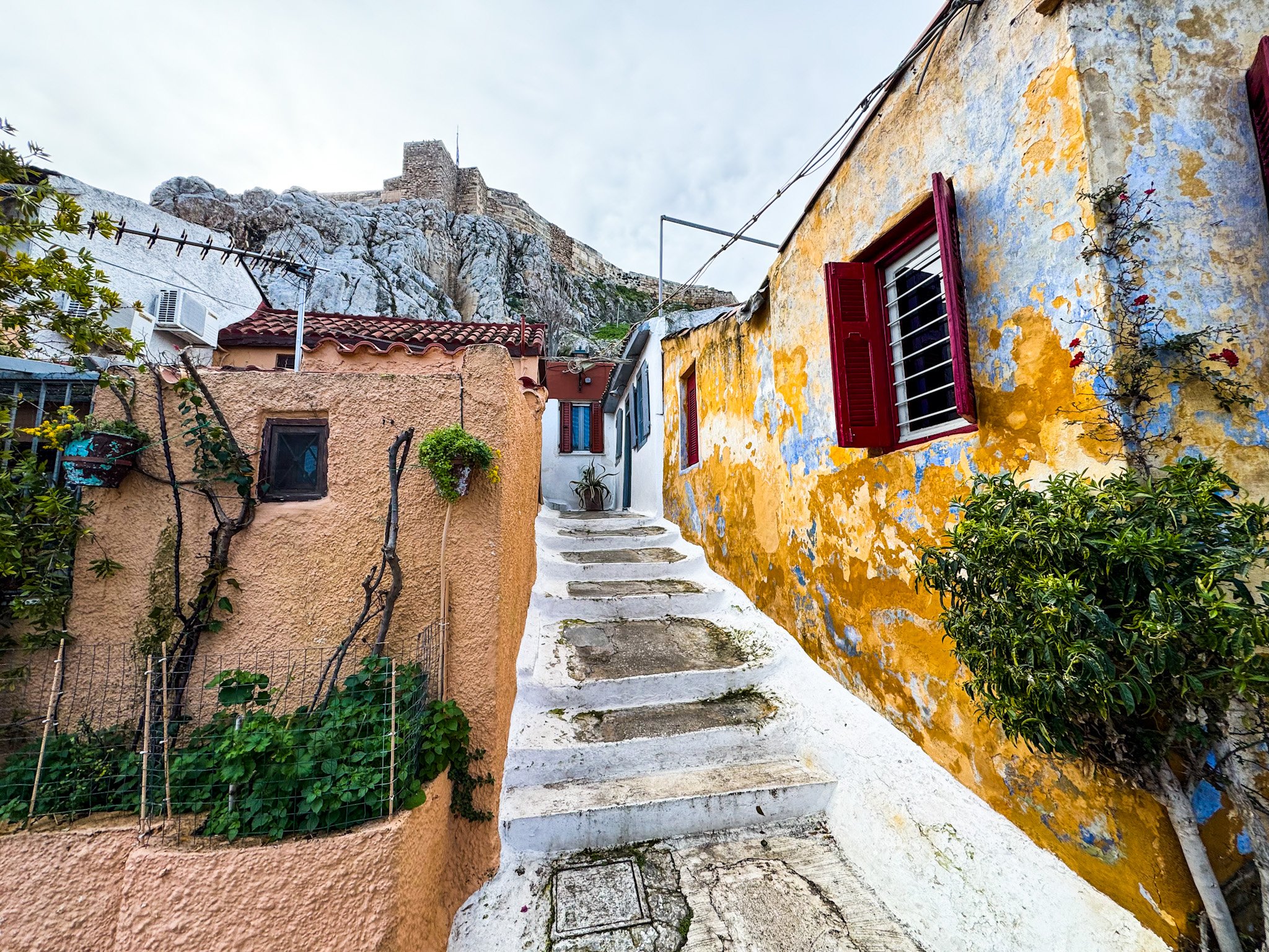 Nietypowy widok na Akropol z wąskiej uliczki w dzielnicy Anafiotika w Atenach. Białe schody prowadzą w górę między kolorowymi domami w stylu cykladzkim, a nad nimi góruje skaliste zbocze i mury obronne Akropolu pod jasnym niebem.