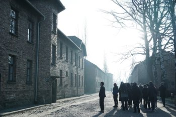 A group of visitors walks down a path at the Auschwitz-Birkenau concentration camp, flanked by old brick buildings under a bright sun.