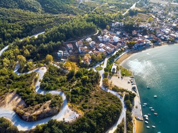 Aerial drone view of a picturesque coastal resort town in Northern Greece (Chalkidiki). A dense green pine forest with a winding mountain road leads down to a typical Greek town with red-tiled roofs. To the right, the sandy beach of Alykes Polygyrou sits beside the turquoise Aegean Sea with a pier and docked boats.
