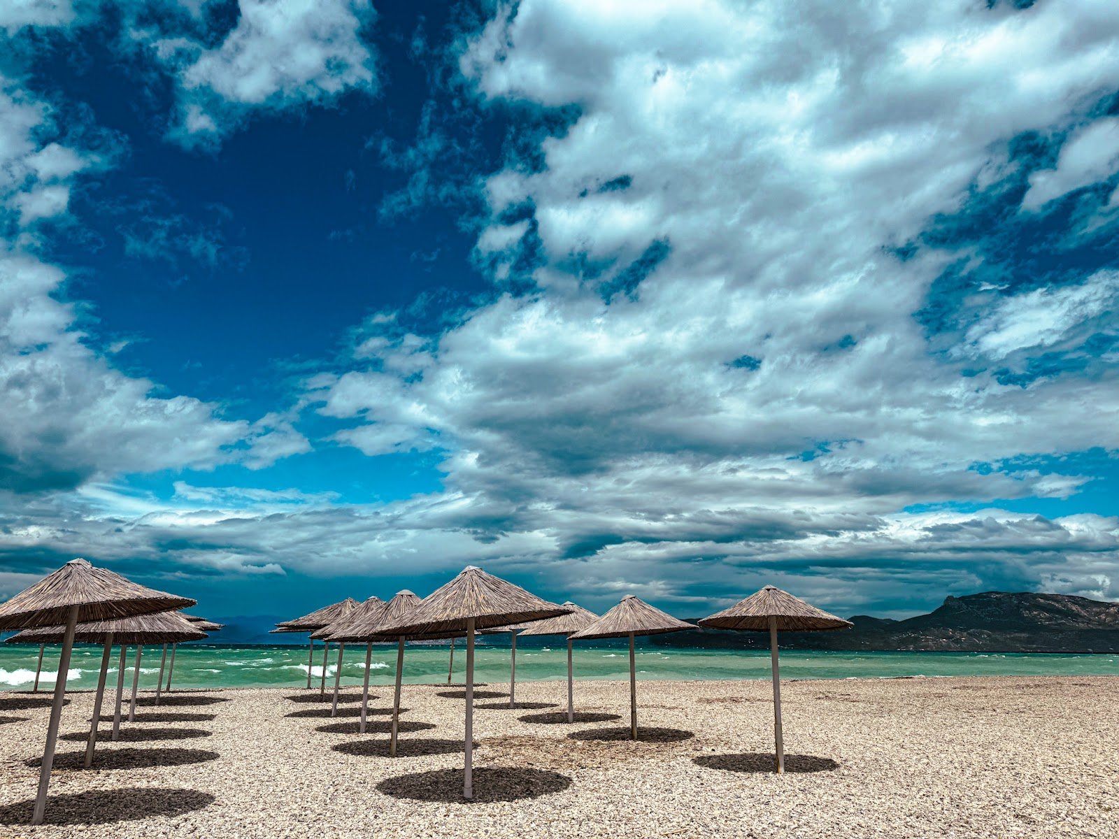 Scenic pebble beach in Corinthia, Greece, featuring straw umbrellas, vibrant turquoise sea, and a mountainous backdrop, highlighting the natural beauty of Peloponnese beaches.