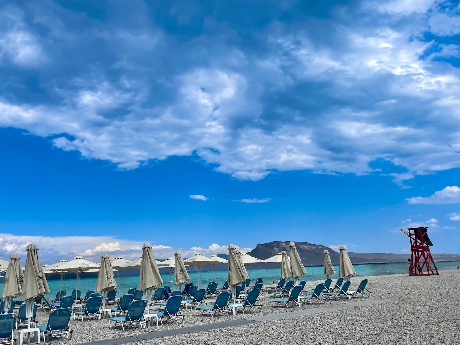 Organized beach with rows of white sunbeds and umbrellas, a red lifeguard tower, and calm turquoise waters in Corinthia, Peloponnese, perfect for a relaxing Athens day trip.